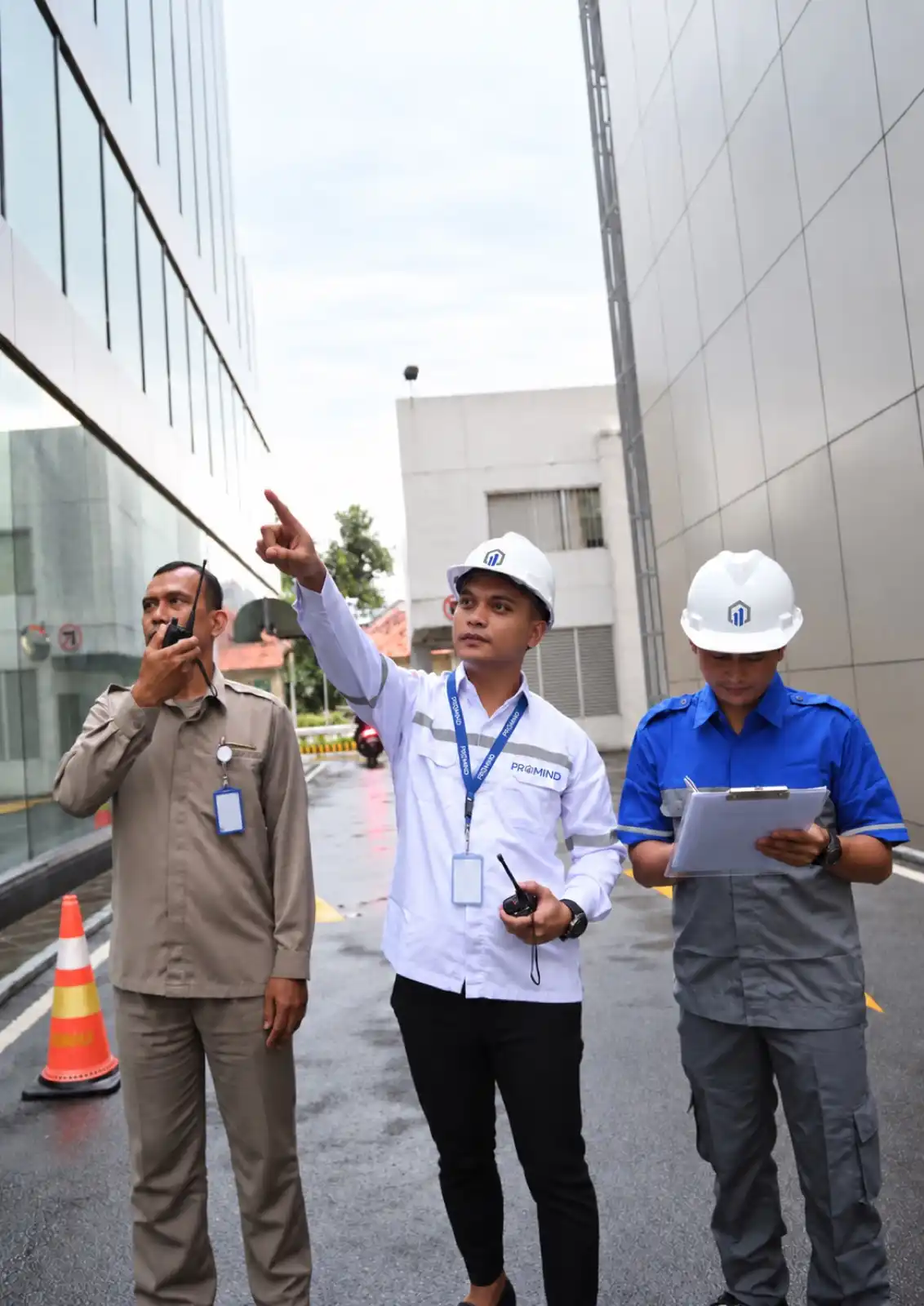 Tiga petugas PROMIND menginspeksi sisi luar gedung: satu berbicara lewat radio, satu menunjuk ke atas, satu mencatat di papan klip.