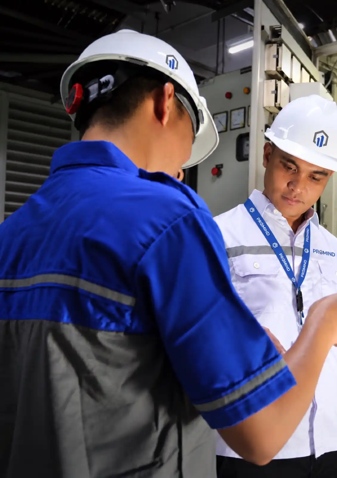 Dua teknisi PROMIND berhelm memeriksa panel kelistrikan di ruang mesin gedung.